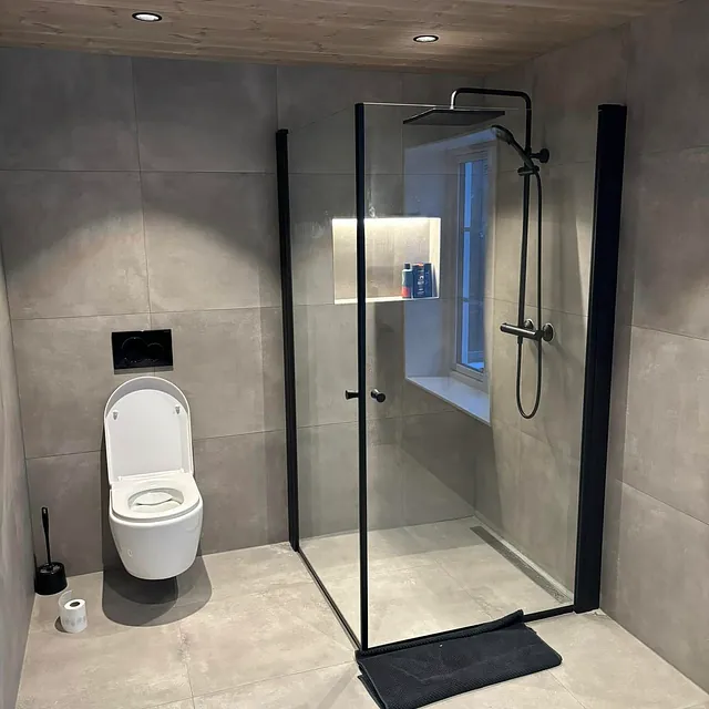 Bad med vegghengt toalett, dusj med glassvegg og grå fliser.