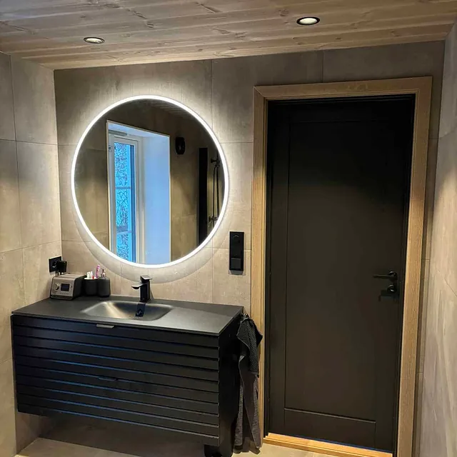 Moderne bad med mørk servant, rund bakbelyst speil og trepanel i taket.