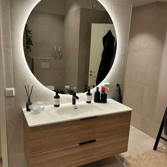 Stort rundt speil med bakbelysning over servant med skuffer i treutførelse.